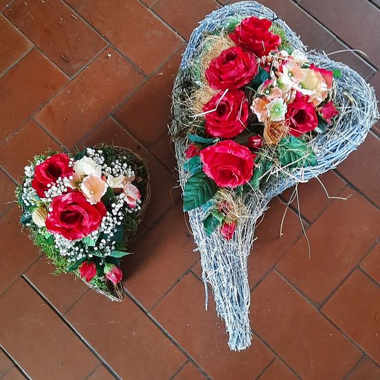 Zwei Blumenarrangements auf einem gefliesten Boden, eines herzförmig und das andere ein größeres Herz mit roten Rosen und anderen Blumen, beide mit getrocknetem Gras und Blättern.