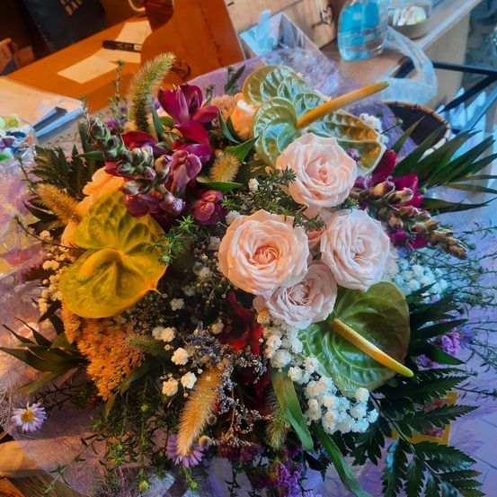 Ein farbenfroher Blumenstrauß mit rosa Rosen, roten Schwertlilien und grünen Blättern, auf einem Tisch arrangiert mit anderen Gegenständen im Hintergrund.