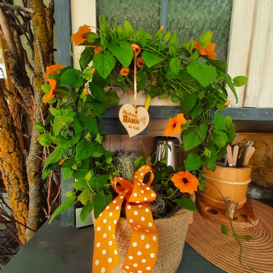 Ein herzförmiger Strauß mit orangefarbenen Blüten in einem geflochtenen Korb, geschmückt mit einem Holzherz, das 'Für die beste Mama' steht. Er ist mit einem orangefarbenen Band gebunden und auf einem Tisch mit anderen Dekorationsartikeln platziert.