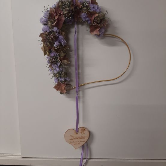 Ein Kranz aus lila Blumen und Grün hängt an einer weißen Wand. Der Kranz ist herzförmig mit einem goldenen Bogen und einem lila Band. Ein holzfarbenes Herzschild mit dem Wort Danke ist am Band befestigt.