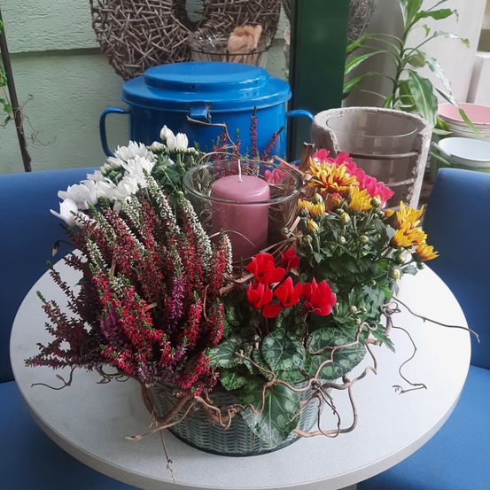 Ein runder Tisch beherbergt eine Blumenarrangement in einem geflochtenen Korb mit roten, weißen und gelben Blumen, begleitet von einer rosa Kerze.