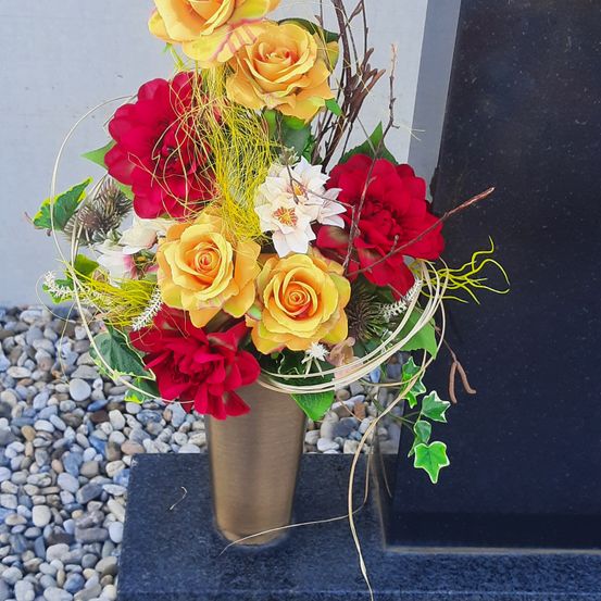 Ein bronzefarbener Vase mit einem farbenfrohen Blumenarrangement aus roten und gelben Rosen, weißen Gänseblümchen und grünen Blättern, auf einer mit Kies bedeckten Granitplatte platziert.