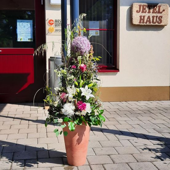 Eine große dekorative Blumenarrangement in einem Terrakotta-Topf steht vor einem Gebäude mit dem Schild 'Jetel Haus'. Die Blumen umfassen weiße, rosa und lila Blüten. Ein Mülleimer befindet sich in der Nähe.