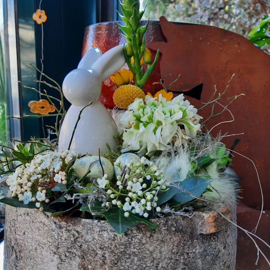 Eine Blumenarrangement in einem Steintopf mit einer weißen Hasenfigur, weißen Blumen und grünen Blättern. Es ist vor dem Hintergrund eines Fensters und eines roten Objekts platziert.