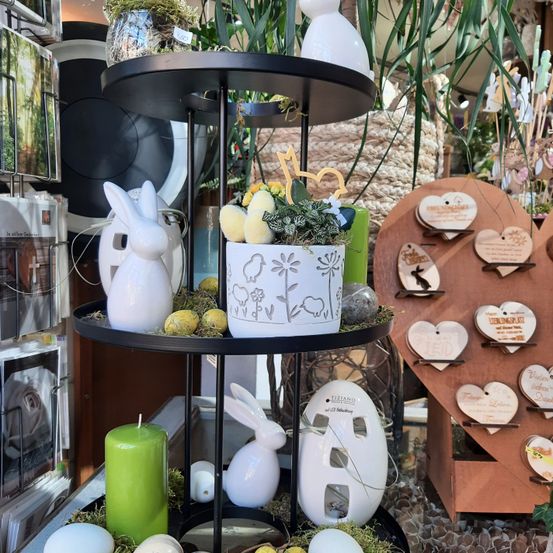 Eine dreistufige Präsentation mit Osterdekorationen, darunter eine grüne Kerze, Keramikhasen und eine Topfpflanze. Im Hintergrund befinden sich herzförmige Holzdekorationen.
