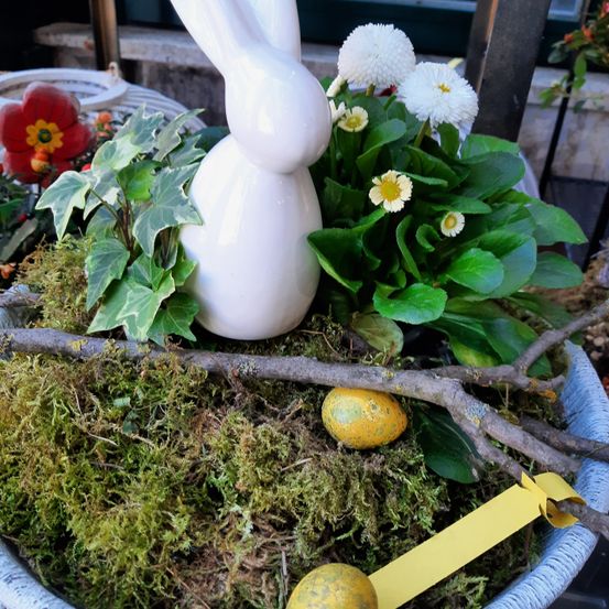 Eine Keramikhasenfigur umgeben von Moos und gelben Eiern, mit weißen Blumen in einem blauen Topf.