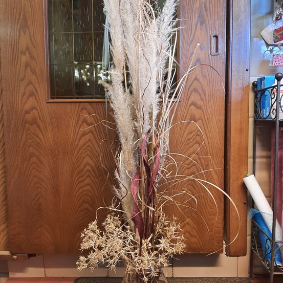Ein hoher Zierpflanzenstrauß mit federigen und spitzen Blättern steht in einem Blumenglas vor einer Holztür mit einem Glasfenster.