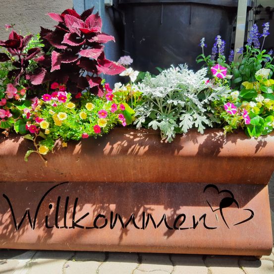 Ein Gartenpflanzer mit einer Vielzahl farbenfroher Blumen, darunter rosa, gelbe und weiße Blüten, vor einer Wand mit einem schwarzen Fass und dem Wort 'Willkommen' darauf geschrieben.