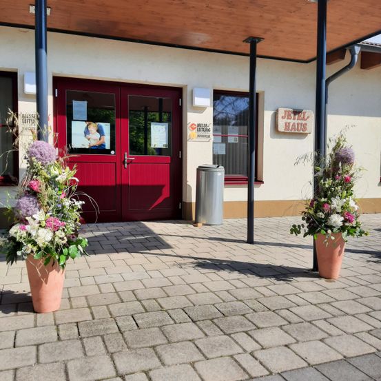Der Eingang eines Gebäudes mit roten Türen und einem Schild, das 'Messe-Lettung' steht. Vor dem Eingang stehen zwei Topfpflanzen. Ein Mülleimer ist neben dem Eingang platziert.