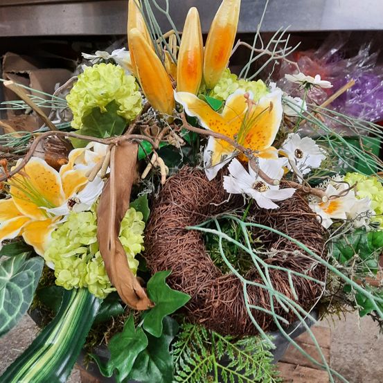 Eine große Blumenarrangement mit einem zentralen getrockneten Nest und verschiedenen Blumen, darunter gelbe Lilien, grüne Hortensien und weiße Blüten. Das Arrangement steht in einem rustikalen Topf.