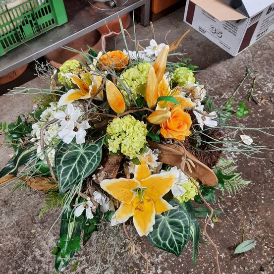 Eine Blumengestecke mit weißen, gelben und grünen Blumen, einschließlich Rosen und Lilien, liegt auf einem Betonboden. Einige Blätter und Stängel sind verstreut.