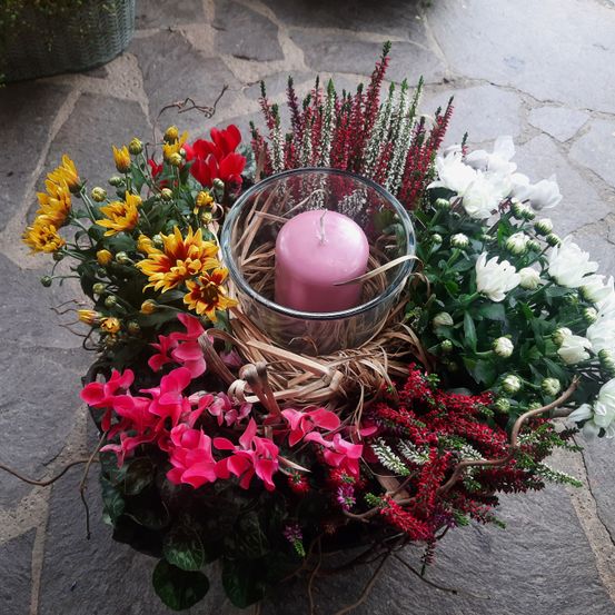 Eine Blumengestecke mit einer rosa Kerze in einem klaren Glasbecher, umgeben von verschiedenen bunten Blumen und Blättern, auf einer Steinoberfläche.