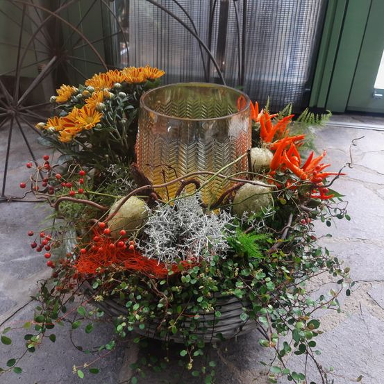 Ein Korb aus Weide enthält eine herbstliche Blumenarrangement mit gelben Blumen, roten Beeren und grünem Laub. In der Mitte steht eine hohe Glasvase, die mit Wasser gefüllt ist.