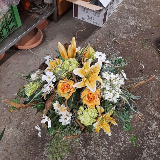 Eine Blumengestecke aus gelben, orangefarbenen und weißen Blumen mit grünen Blättern auf einem Betonboden, neben einem Lagerbox und einem Regal.