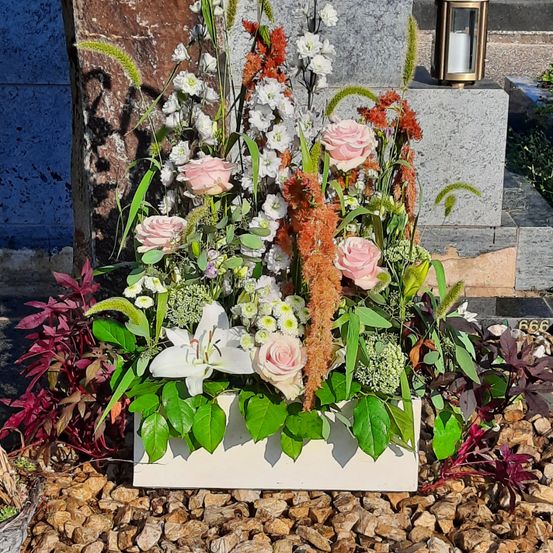 Eine weiße Blumenarrangement mit rosa und weißen Rosen, weißen Lilien und grünen Blättern in einer weißen Box auf einer felsigen Oberfläche.