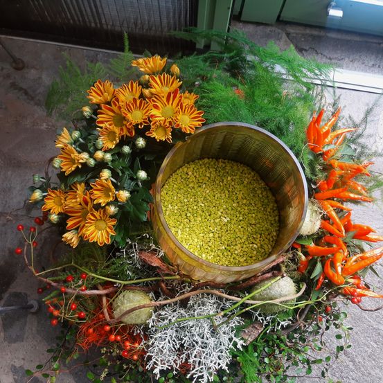 Ein kreisförmiges Blumenarrangement mit gelben Gänseblümchen, roten Beeren und grünen Blättern. In der Mitte steht eine Schüssel mit grünen Erbsen zwischen den Blumen.