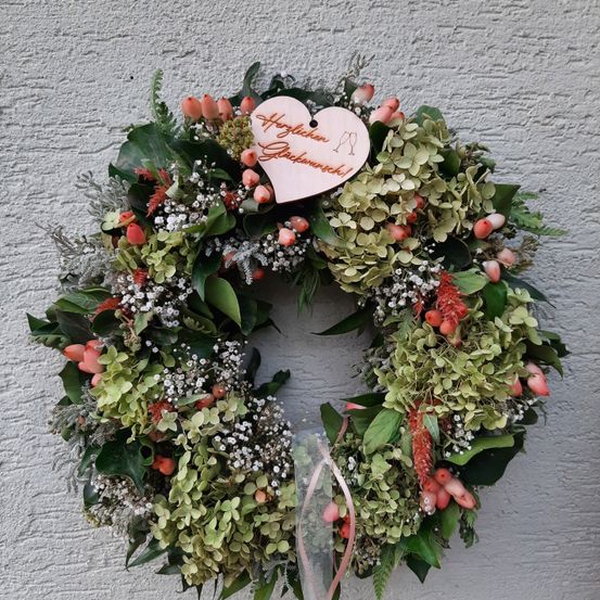 Ein Kranz aus grünen Blättern und Blumen hängt an einer Wand. Der Kranz hat ein herzförmiges Schild, auf dem Herzlichen Glückwunsch! steht. Der Kranz ist mit einer Schleife verziert.