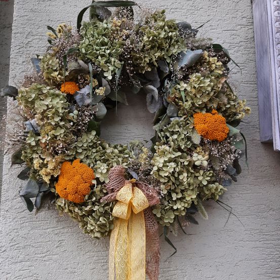 Ein Kranz aus getrockneten Blumen und Blättern hängt an einer Wand, mit einem gelben Band und orangefarbenen Blumen als Highlights.