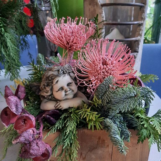 Ein hölzerner Pflanzkasten mit einer Cherub-Figur und rosa Blumen, umgeben von grünen Blättern und lila Blüten, auf einem Tisch.