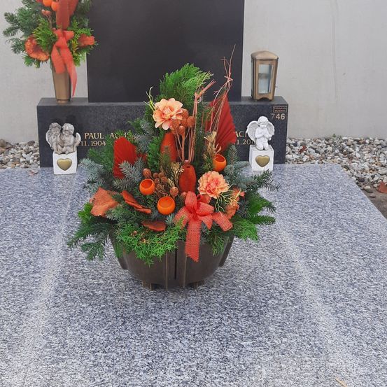 Ein Blumenarrangement mit orangen und roten Blumen in einem braunen Topf steht auf einer grauen Oberfläche vor einem Grabstein für Paul.