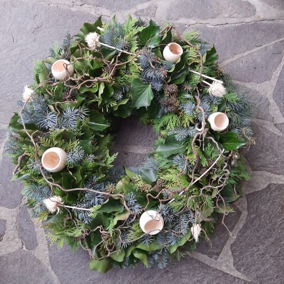 Ein Kranz aus grünen Blättern, Tannenzapfen und Zweigen liegt auf einer Steinoberfläche. Es gibt sechs weiße Kerzen auf dem Kranz.