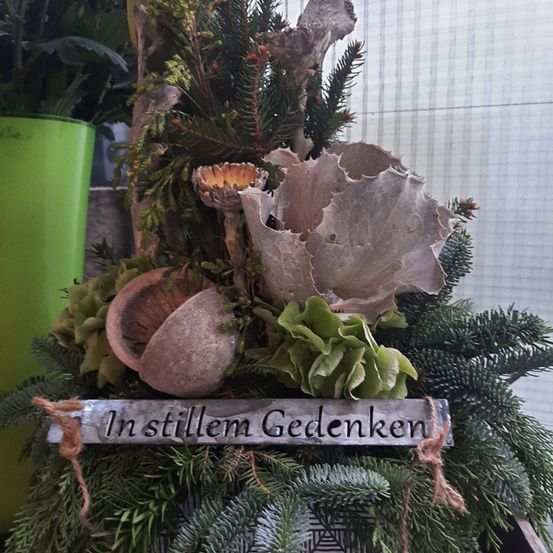Eine Blumenarrangement mit einem Metallschild, das 'In stillem Gedenken' sagt, ist vor einem grünen Topf und einem Fenster platziert.