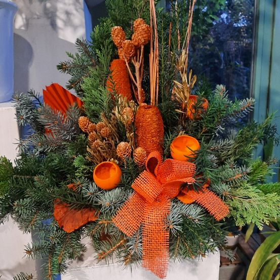 Eine farbenfrohe Blumenkomposition mit leuchtend orangefarbenen Akzenten und grünen Kiefernzweigen, gebunden mit einem orangefarbenen Band, vor einem unscharfen Fensterhintergrund.