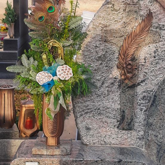 Ein Blumenarrangement in einem bronzefarbenen Vasen steht auf einer Granitoberfläche in der Nähe einer großen Steinmauer. Das Arrangement besteht aus grünen Blättern, blauen Bändern und einem Pfauenfeder. Ein bronzefarbener Lampenschirm und eine weitere Pflanze sind in der Nähe.