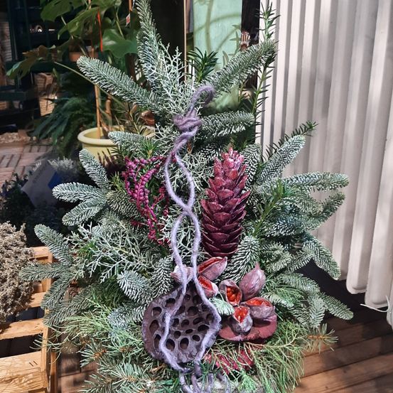 Ein dekorierter Weihnachtsbaum mit lila und roten Ornamenten ist in einem Raum ausgestellt. Er hat Nadeln und eine lilafarbene Ranke. Dahinter befinden sich einige Pflanzen und ein weißer Vorhang.