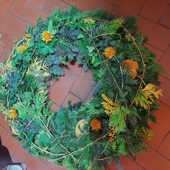 Ein kreisförmiger Kranz aus verschiedenen immergrünen Pflanzen und Zweigen, geschmückt mit gelben Blumen, liegt auf einem roten Fliesenboden.