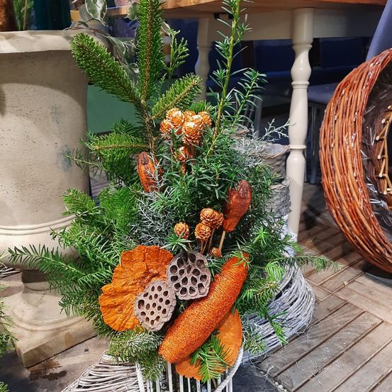 Ein Nahaufnahme einer dekorativen Blumenarrangement mit orangen und grünen Blättern, Zapfen und Zweigen, präsentiert in einem geflochtenen Korb auf einem Holzboden.