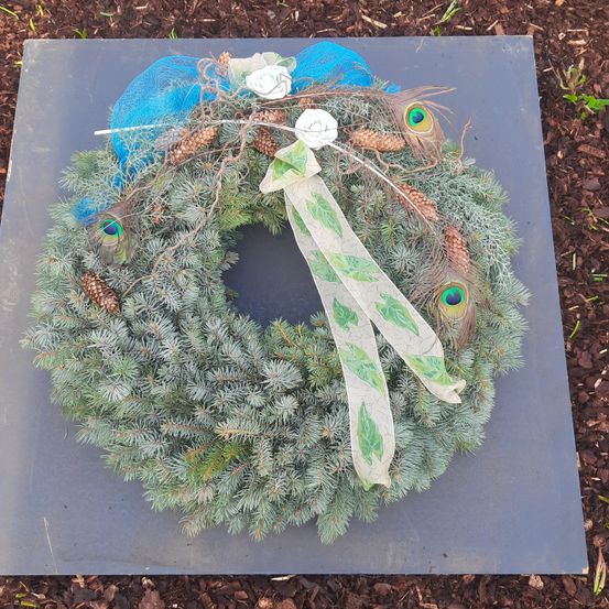 Ein Kranz aus immergrünen Zweigen, verziert mit blauen Bändern, Pfauenfedern und Tannenzapfen, liegt auf einer grauen Oberfläche.