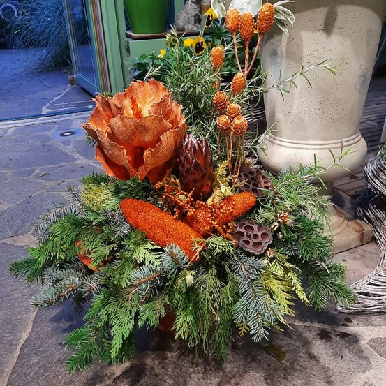 Eine Blumenarrangement mit orangen und grünen Blättern, einem Zapfen und Beeren liegt vor einem Gebäude auf dem Boden.