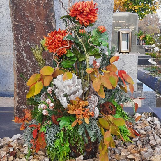 Eine farbenfrohe Blumendekoration mit Herbstblättern, orangefarbenen Blumen und Tannenzapfen, die vor einem grauen Grabstein mit dem Namen Beck auf Felsen platziert ist.