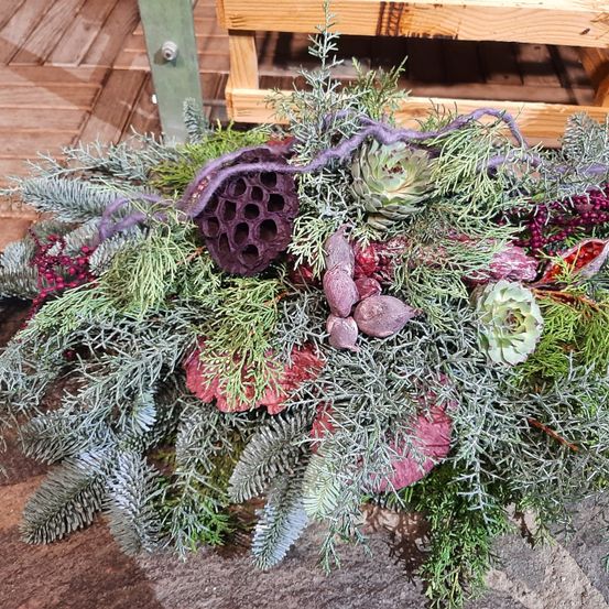 Eine dekorative Blumenarrangement mit silbernen, roten und lila Blättern, Sukkulenten und einer lilafarbenen Honigwabenstruktur auf einer Holzoberfläche.