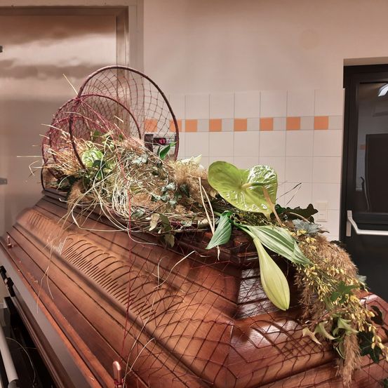 Ein hölzerner Sarg ist mit verschiedenen Pflanzen und Gräsern dekoriert. Er hat einen Netzbezug, der mit mehreren roten Netzen verziert ist. Der Sarg steht an einer gefliesten Wand, mit einer Tür auf der rechten Seite.