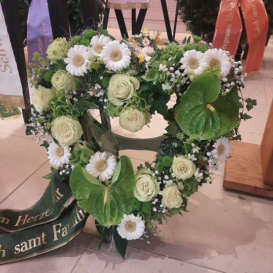 Ein herzförmiges Blumenarrangement mit weißen und grünen Blumen, einschließlich Rosen und Babys Atmen, steht auf einem gefliesten Boden. Es hat ein grünes Band mit dem Text 'sammt Familie'. Dahinter befinden sich einige Banner.
