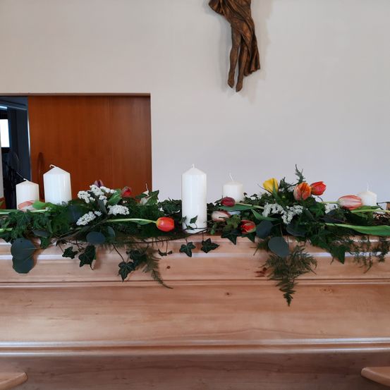 Ein hölzerner Sarg ist mit weißen und roten Blumen, grünen Blättern und fünf weißen Kerzen auf einer Bühne geschmückt. Dahinter ist eine Holztür und ein Wandkreuz zu sehen.