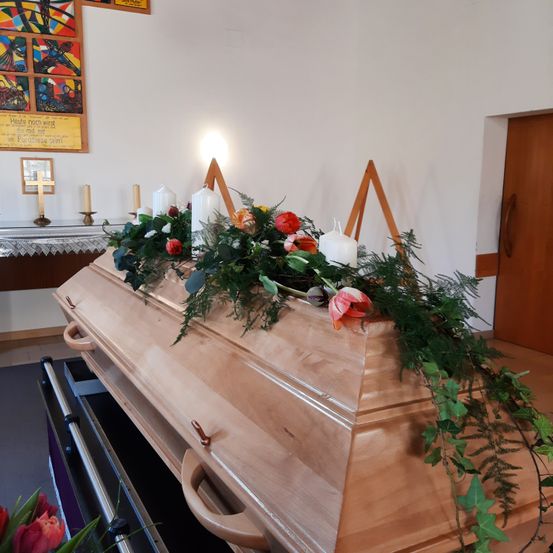 Ein hölzerner Sarg ist mit Blumen und Kerzen geschmückt. Er steht auf einer Plattform mit Griffen. Dahinter befindet sich eine weiße Wand mit einem gerahmten Bild und zwei brennenden Kerzen.