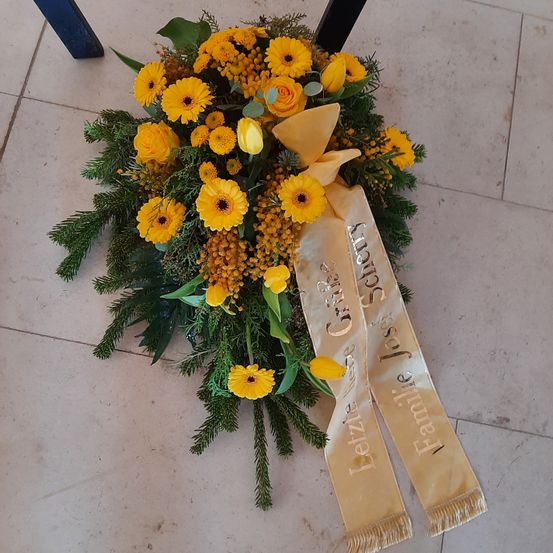 Ein gelbes Blumenarrangement mit grünen Blättern liegt auf einem weißen gefliesten Boden. Es hat ein Band mit dem Text 'Famille Jose et Lucie' und 'Grises Lerze'.