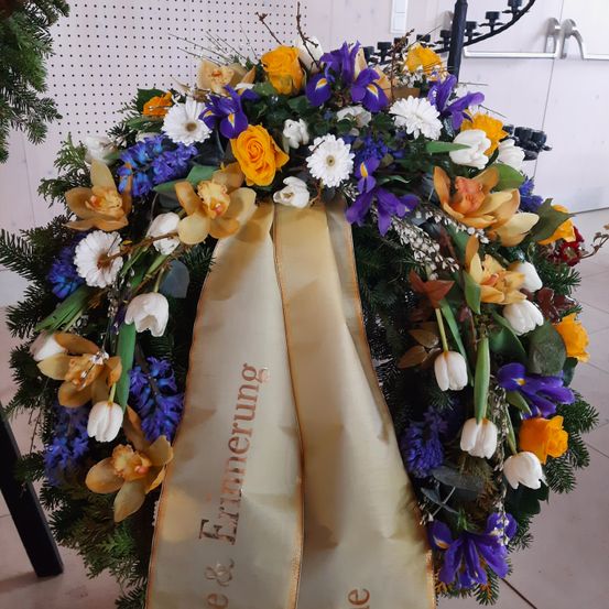 Ein Blumenkranz mit weißen, gelben, blauen und lilafarbenen Blumen. Der Kranz hat ein goldenes Band mit dem Text Erinnerung. Hinter dem Kranz befindet sich eine weiße Wand mit einem schwarzen Metallobjekt.