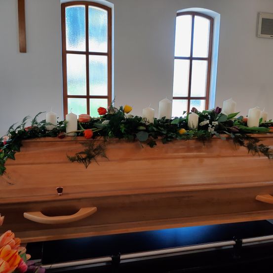 Ein hölzerner Sarg mit Blumenarrangement und Kerzen auf einem Ständer in einem Raum mit weißen Wänden und bogenförmigen Fenstern.