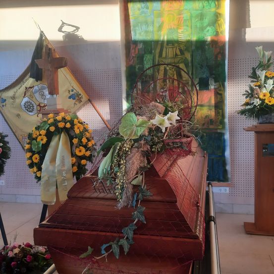 Ein hölzerner Sarg ist mit Blumen und grünen Blättern verziert. Dahinter ist eine Flagge mit einem Kreuz und einem Kranz zu sehen. Rechts hängt ein farbenfrohes Gemälde an der Wand.