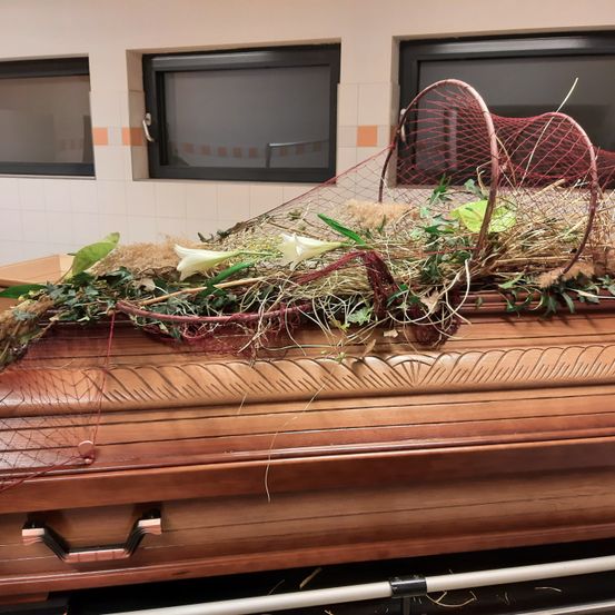 Ein hölzerner Sarg mit komplizierten Mustern ruht auf einem Metallwagen, geschmückt mit einem Netz, Blumen und Grünpflanzen. Hinter ihm sind drei Fenster mit schwarzen Rahmen zu sehen.