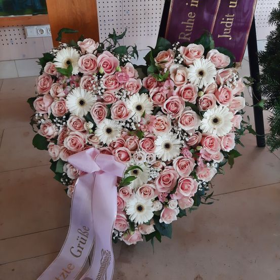 Ein herzförmiges Blumenarrangement aus rosa und weißen Rosen und Gänseblümchen, verziert mit einer rosa Schleife und grünen Blättern, auf einem gefliesten Boden platziert.