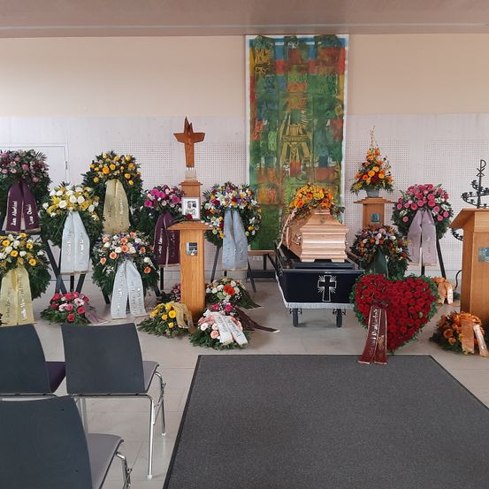 Ein Trauerfeier-Setting mit einem Kreuz, Blumenarrangements und einem Sarg auf einem Wagen. Stühle sind in Reihen angeordnet und ein Holzpodium ist vorhanden.