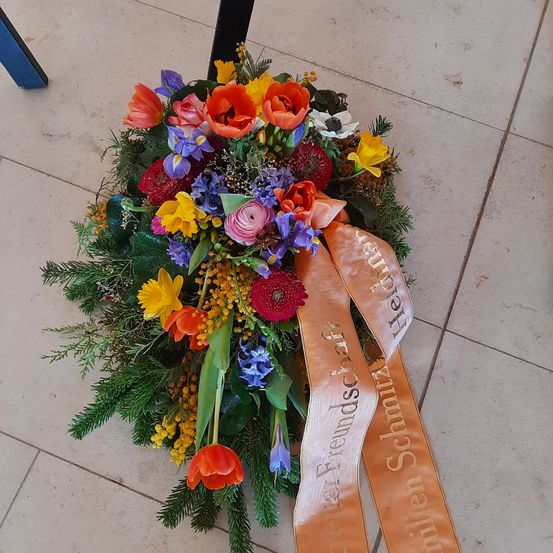 Ein farbenfroher Blumenstrauß mit verschiedenen Blumen, darunter Rosen, Tulpen und Narzissen, liegt auf einem Fliesenboden. Er hat ein orangefarbenes Band mit goldenem Text, der 'Herzlichen Dank' lautet.