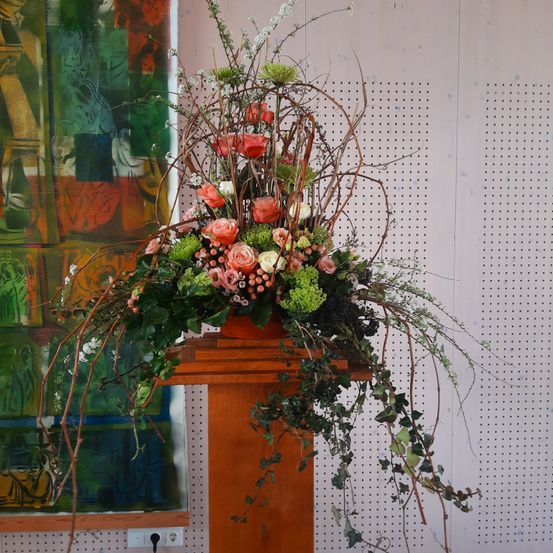 Eine elegante Blumenkomposition mit rosa, weißen und roten Blumen sowie grünem Laub steht auf einem Holzpodest. Die Blumen sind in einem Topf angeordnet und von Ästen umgeben. Der Hintergrund ist eine weiße Wand mit einigen Löchern.