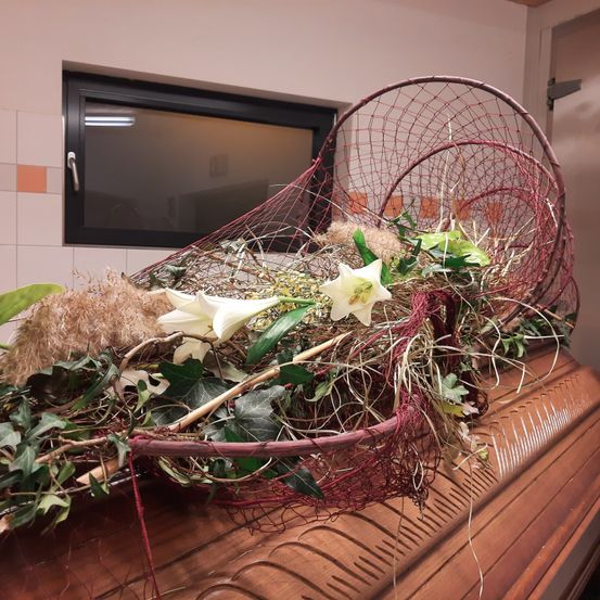 Ein Sarg ist mit einer Blumendekoration in einem roten Netz geschmückt und befindet sich in einem Raum mit weiß gefliester Wand und Glasfenster.