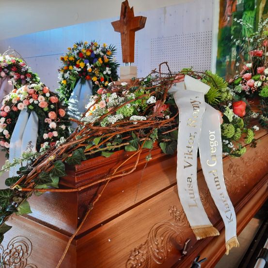 Ein hölzerner Sarg, geschmückt mit Blumenarrangements und einem dekorativen Kranz, mit einem weißen Band, auf dem 'Luisa Viktor und Klara Gregor' steht. Ein Kreuz und zusätzliche Blumenkränze im Hintergrund.